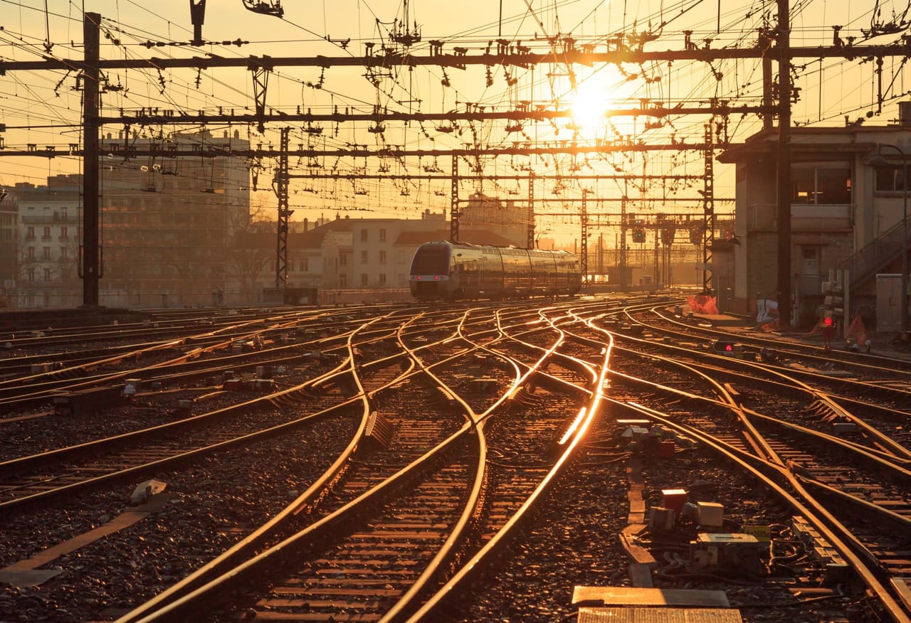 Faisceau de voies ferrées et caténaires au soleil levant, un train à l’arrière-plan